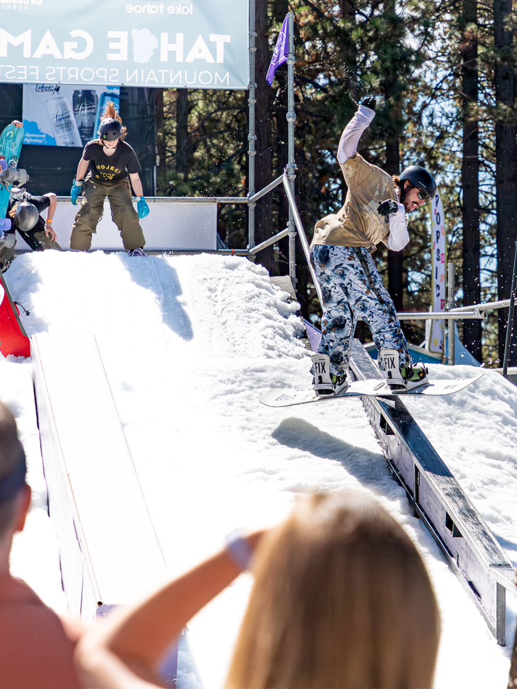 A young man dressed in summer clothes snowboards down a ramp, while other competitors watch him perform.