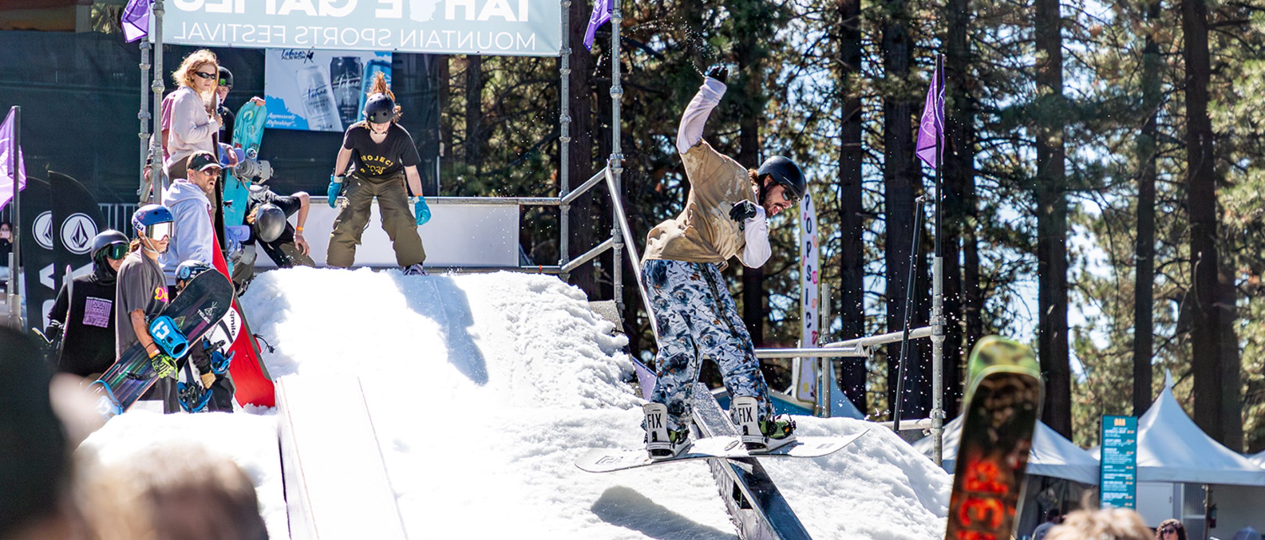 A young man dressed in summer clothes snowboards down a ramp, while other competitors watch him perform.